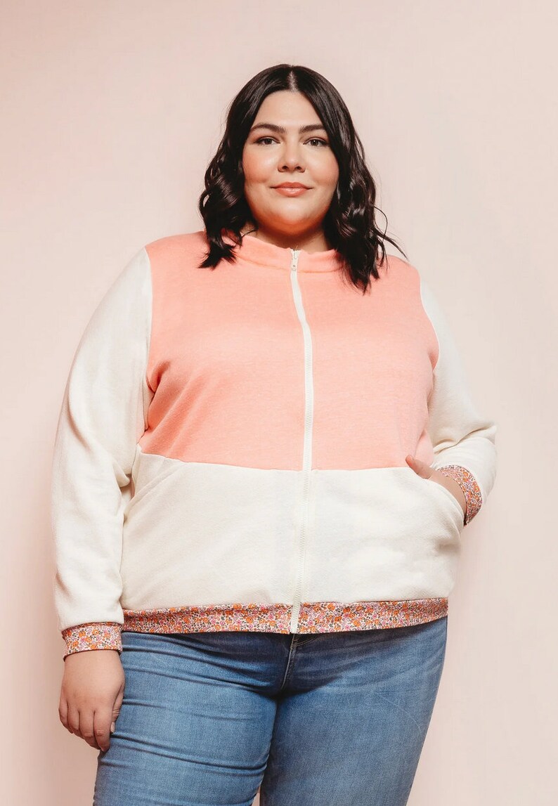 Puede incluir: Una mujer lleva una chaqueta con cremallera rosa y blanca con un ribete de estampado floral. La chaqueta tiene una cremallera blanca y un bolsillo de parche en el lado izquierdo. Lleva pantalones vaqueros azules.