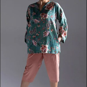 May include: A woman wearing a teal floral print top with a v-neck and long sleeves. She is also wearing pink cropped trousers and green sandals.