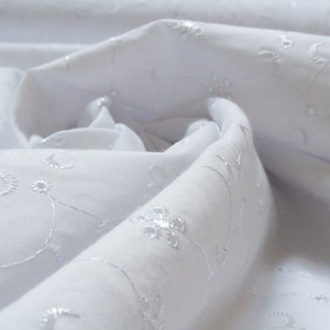 May include: White fabric with a delicate embroidered floral pattern. The fabric is soft and flowing, creating a romantic and elegant look.