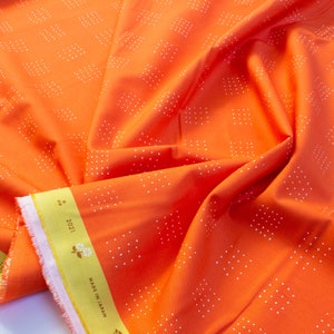 May include: Bright orange fabric with a repeating pattern of small white squares. The fabric is folded, showing its texture and drape. A yellow label with text and floral design is visible along the edge. The fabric is made in Japan.