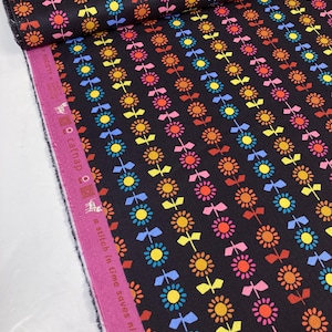 May include: A roll of black fabric featuring a repeating pattern of colourful flowers and bows. The flowers are in yellow, orange, pink, and blue. The fabric has a pink selvage with text.