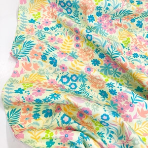 May include: Floral fabric with a cream background, featuring a pattern of pink, blue, yellow, and green flowers and foliage. The fabric is soft and flowing, with visible folds and creases. The design includes various flower types and leaf shapes.