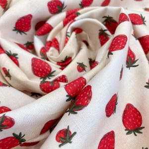 May include: Close-up of a cream fabric featuring a repeating pattern of red strawberries. Each strawberry has green leaves. The fabric is folded, creating shadows and depth, with the strawberries scattered across it.