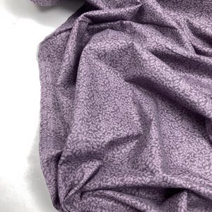 May include: A close-up of a fabric with a floral pattern. The fabric is a muted purple colour with a repeating pattern of small flowers and leaves in a lighter shade of purple. The fabric is draped, showing folds and texture.
