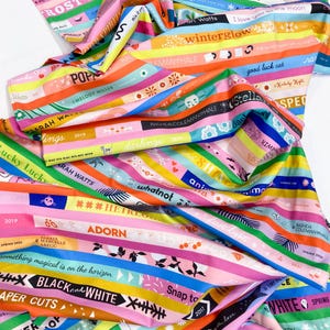 May include: A vibrant fabric with colorful horizontal stripes, each bearing text in various fonts and colors. The fabric features words like "winterglow," "darling," and "black and white." The design includes floral and geometric patterns, creating a lively and artistic textile.