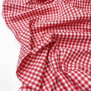 May include: Red and white gingham fabric with a checkered pattern. The fabric is folded and draped, showing the texture and weave.