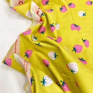 May include: Yellow fabric with a repeating pattern of pink and white strawberries. The fabric is folded and the text "@knelkigh 100% Cotton Made in Japan" is visible on the side.