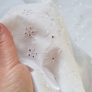 May include: White cotton fabric with a delicate eyelet embroidery pattern. The fabric is folded and shows the intricate design of small flowers and circles.
