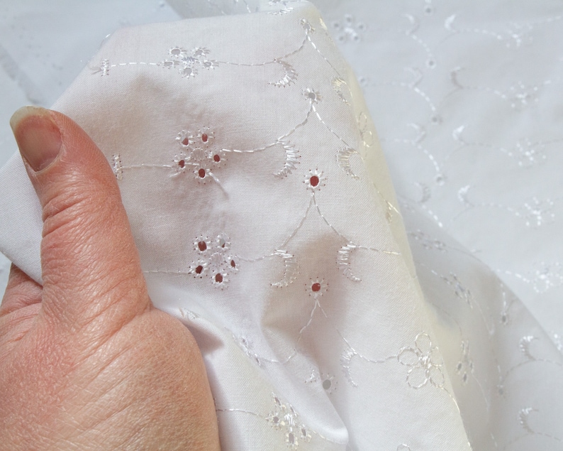 May include: White cotton fabric with a delicate eyelet embroidery pattern. The fabric is folded and shows the intricate design of small flowers and circles.