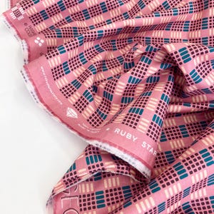 May include: Pink fabric featuring a geometric design of squares and rectangles in teal, white, and dark purple. The fabric is labelled "RUBY STAR" and "100% Cotton Made in Japan". The fabric is folded and draped.