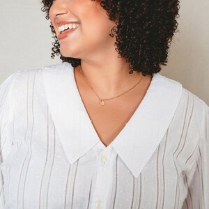 May include: A woman with curly brown hair is smiling and wearing a white button-down shirt with a white collar and thin vertical stripes. She is wearing a gold necklace with a small round pendant.
