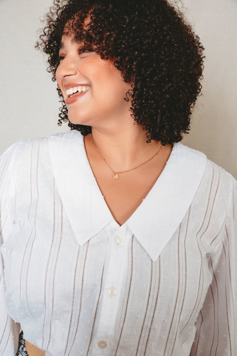 May include: A woman with curly brown hair is smiling and wearing a white button-down shirt with a white collar and thin vertical stripes. She is wearing a gold necklace with a small round pendant.