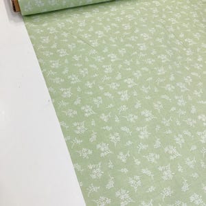 May include: A roll of pale green fabric featuring a repeating pattern of tiny white floral sprigs. The fabric is presented on a white surface, with a section of the roll visible, highlighting the texture and design.