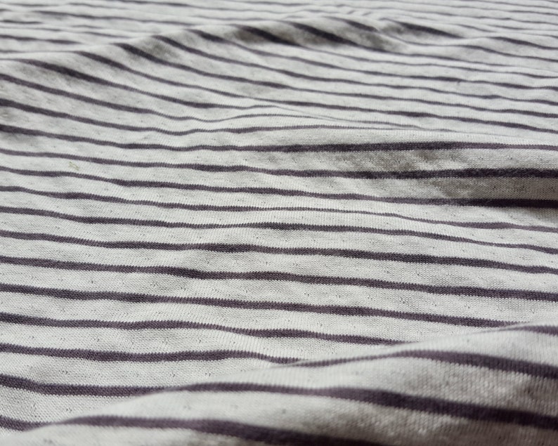 May include: Close-up of a white and gray striped fabric with a subtle texture. The fabric is wrinkled and has a soft, casual look.