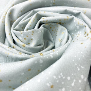 May include: Close-up of a fabric with a pale blue-green hue, speckled with white and gold-coloured dots. The fabric is tightly rolled, showcasing its texture and pattern. Suitable for sewing, quilting, or craft projects.