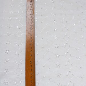May include: White cotton fabric with a repeating pattern of small white embroidered flowers and curved lines. A wooden ruler with inch markings is placed along the left edge of the fabric.