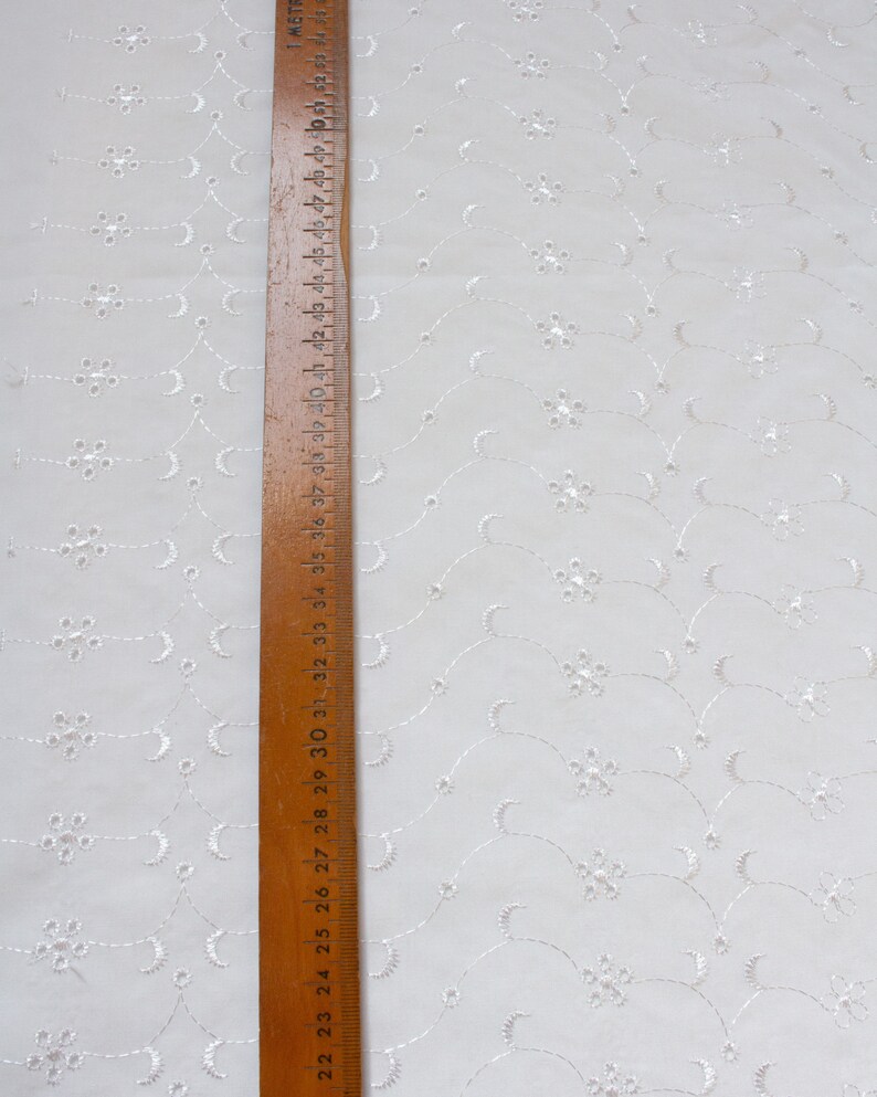 May include: White cotton fabric with a repeating pattern of small white embroidered flowers and curved lines. A wooden ruler with inch markings is placed along the left edge of the fabric.
