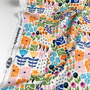 以下が含まれることがあります： 白地にカラフルな花柄の生地。オレンジ、青、ピンク、黄色の花と緑の葉と茎がデザインされています。生地には「CUR8TOR Modernista」と「AGF Studio」の文字がプリントされています。