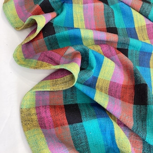 May include: A colourful plaid fabric with a woven texture. The pattern features large squares in shades of red, pink, yellow, teal, blue, and black. The fabric is draped, showing the texture and colour variations.