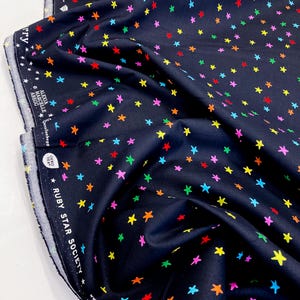 May include: Navy blue fabric featuring a random pattern of multi-coloured stars. The fabric is printed with stars in red, yellow, green, blue, and pink. The fabric is labelled "Ruby Star Society" and is 100% cotton.