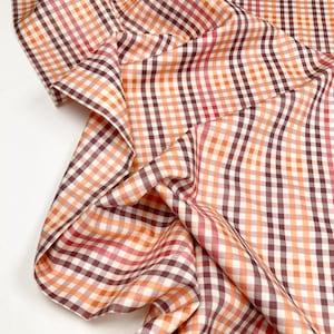 May include: Close-up of a fabric with a classic gingham pattern. The fabric features a repeating checkered design in shades of white, orange, red, and brown. The fabric is folded and draped, showcasing its texture and pattern.