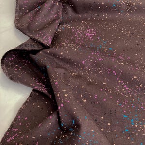 May include: Dark brown fabric with a speckled pattern of pink, blue, and beige dots. The fabric is folded, showing the texture and the raw edge of the material. The fabric is likely used for sewing or crafting projects.