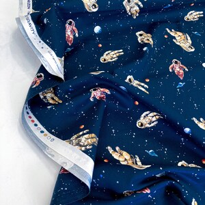 May include: Dark blue fabric featuring a repeating pattern of astronauts, planets, and stars. The fabric has a white border with the text "GRAVITY" and "BEYOND THE HORIZON". Suitable for quilting or other craft projects.