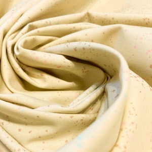 May include: A close-up of a soft, beige fabric with a subtle speckled pattern in shades of pink, blue, and brown. The fabric is folded and draped, showcasing its texture and the delicate pattern.