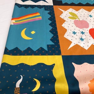 May include: A colorful fabric with a patchwork design featuring celestial and nature-inspired motifs. The pattern includes rainbows, moons, stars, and stylized floral elements in shades of blue, yellow, orange, and white. The fabric is folded, showing the repeating pattern.