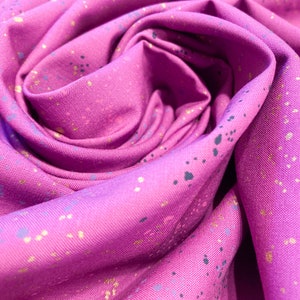 以下が含まれることがあります： ピンクの生地のクローズアップ。金と黒の斑点模様が散りばめられています。生地は折り畳まれ、しわくちゃになっており、質感と模様がわかります。