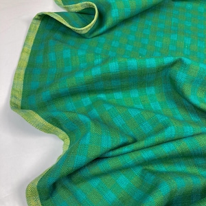 May include: A folded green and teal checkered fabric with a yellow-green border. The fabric has a woven texture and is draped, showing the pattern and colour variations. The edges are neatly finished.
