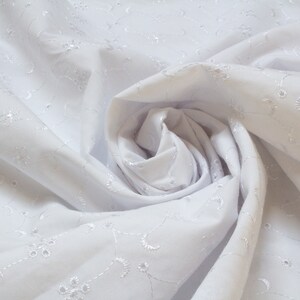 May include: White cotton fabric with a delicate embroidered floral pattern. The fabric is gathered and folded to create a soft, flowing effect.