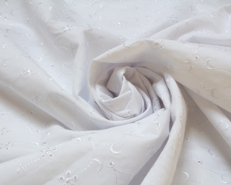 May include: White cotton fabric with a delicate embroidered floral pattern. The fabric is gathered and folded to create a soft, flowing effect.