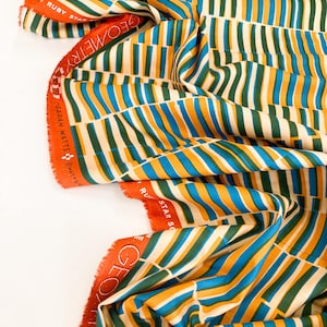 May include: A folded fabric featuring a repeating vertical stripe pattern in teal, green, yellow, and off-white. The selvage edge is orange with the text "RUBY STAR SOCIETY" and "GEOMETRY". The fabric is likely cotton or a similar material.