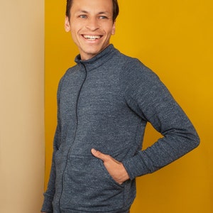 Puede incluir: Un hombre con un su&eacute;ter azul con cremallera y una mano en el bolsillo, de pie frente a un fondo amarillo.