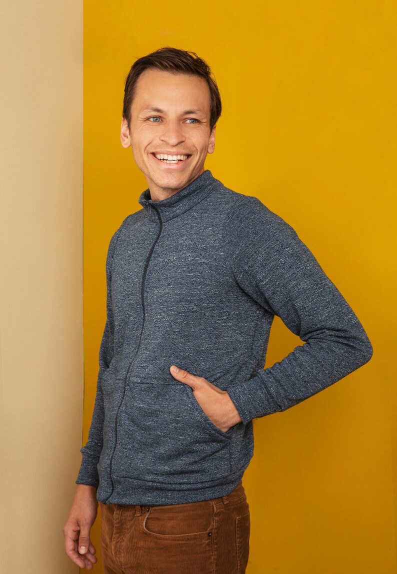 Puede incluir: Un hombre con un su&eacute;ter azul con cremallera y una mano en el bolsillo, de pie frente a un fondo amarillo.