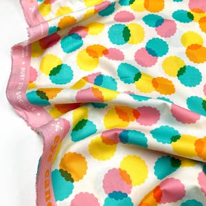 May include: White fabric with a repeating pattern of colourful, overlapping circles. The circles are in shades of pink, yellow, orange, green, and blue. The fabric has an "eye candy" label on the edge.