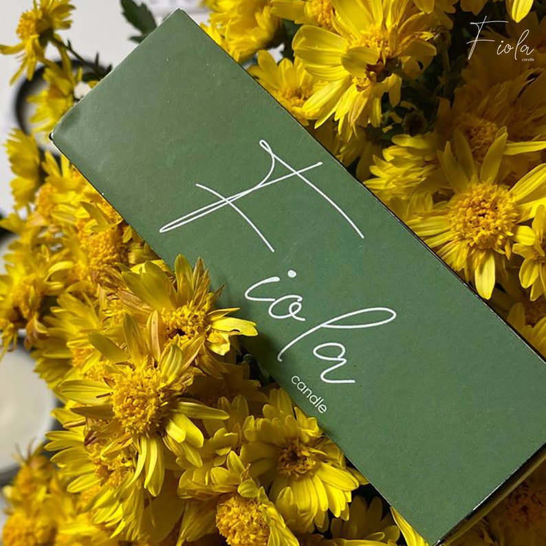 May include: A green candle box with the text "Fioia candle" in white script. The box is surrounded by yellow flowers.