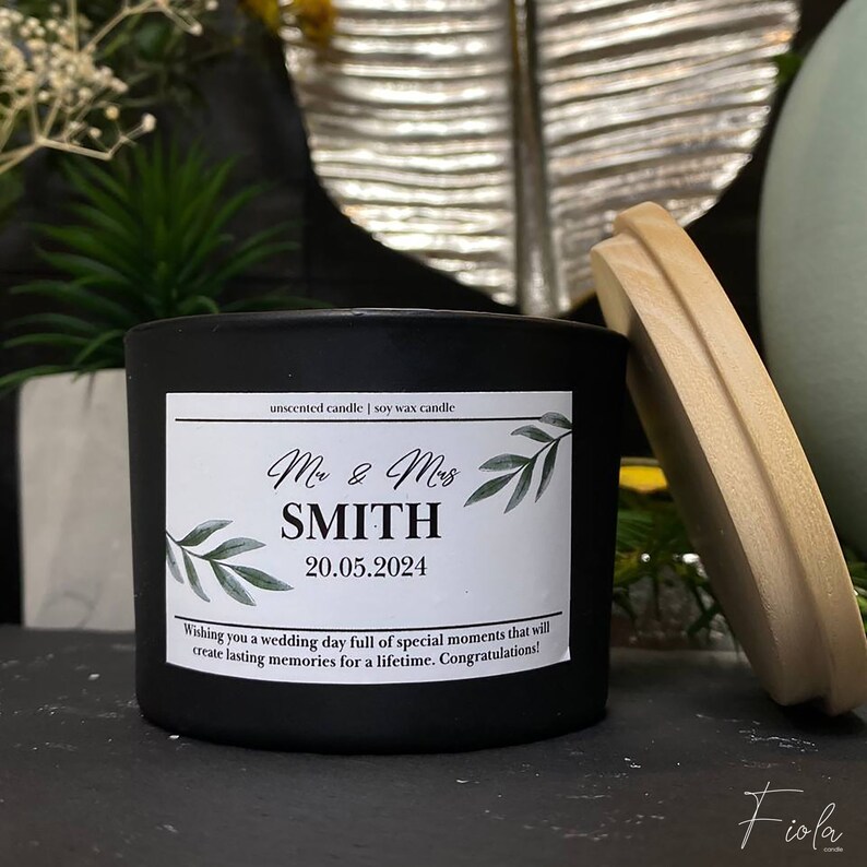 May include: Black soy wax candle with a white label that says "unscented candle | soy wax candle", "Mr. & Mrs. SMITH", "20.05.2024", and "Wishing you a wedding day full of special moments that will create lasting memories for a lifetime. Congratulations!" The label has a green leafy design.