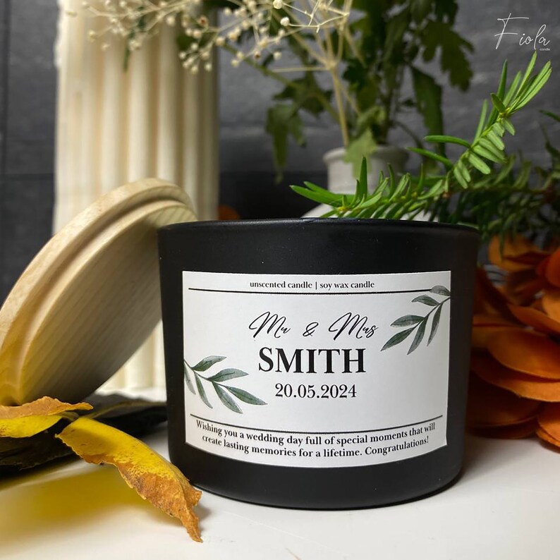 May include: Black soy wax candle with a white label that says "unscented candle | soy wax candle". The label also has a green leafy design and the text "Mr & Mrs SMITH 20.05.2024 Wishing you a wedding day full of special moments that will create lasting memories for a lifetime. Congratulations!"