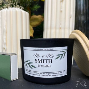 May include: Black soy wax candle with a white label that says "unscented candle | soy wax candle" and features a wedding design with green leaves and the text "Mr & Mrs SMITH 20.05.2024 Wishing you a wedding day full of special moments that will create lasting memories for a lifetime. Congratulations!"