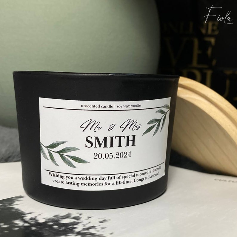 May include: Black soy wax candle with a white label. The label has green leaves and the text "Mr & Mrs SMITH 20.05.2024" and "Wishing you a wedding day full of special moments that will create lasting memories for a lifetime. Congratulations!"