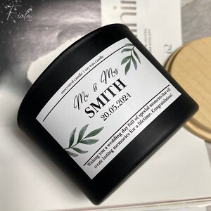 May include: Black soy wax candle with a white label. The label has a green leafy design and the text "Mr & Mrs SMITH 20.05.2024 Wishing you a wedding day full of special moments that will create lasting memories for a lifetime. Congratulations!"