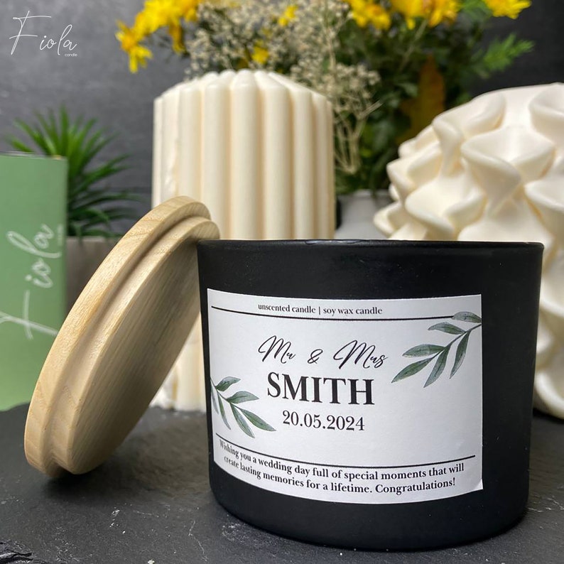 May include: Black soy wax candle with a white label that says "unscented candle | soy wax candle" and "Mr & Mrs SMITH 20.05.2024". The label also has a green leafy design. The candle has a wooden lid.