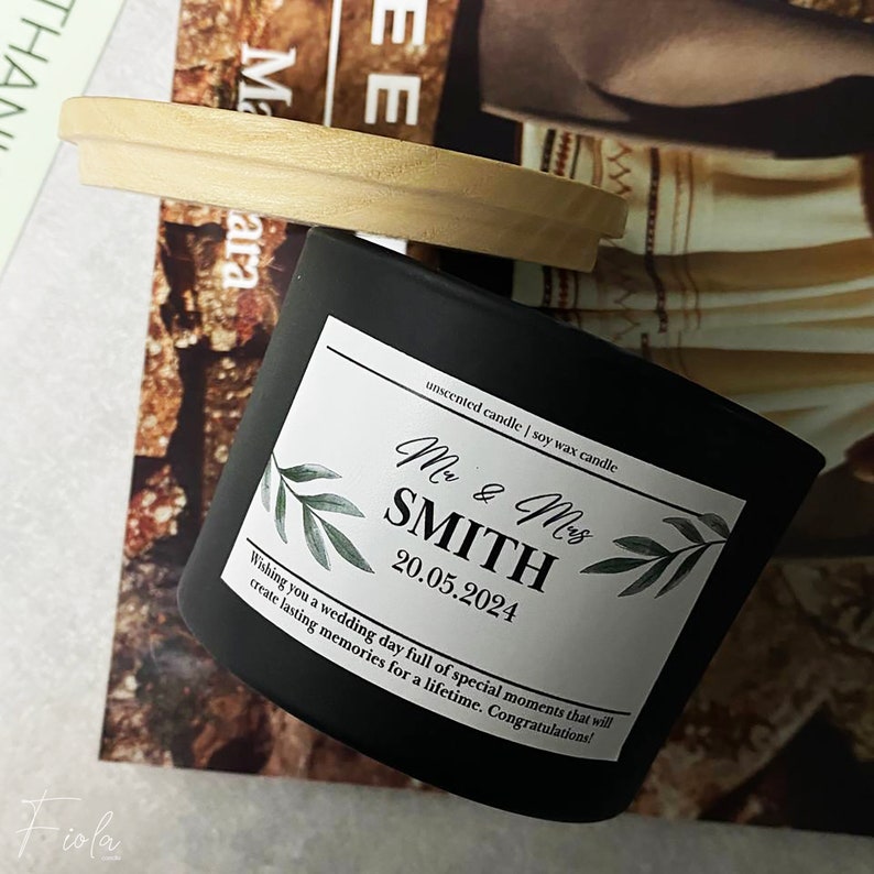 May include: Black soy wax candle with a white label that says "Mr. & Mrs. SMITH 20.05.2024" and "unscented candle | soy wax candle". The label also has a green leafy design. The candle is on a wooden lid.