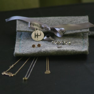 Op de afbeelding: Een grijze wollen tas met een zilveren hanger en een zilveren ketting. De tas is vastgebonden met een zilveren lint en heeft het woord "Velgda" erop gedrukt. De tas ligt op een zwart oppervlak.