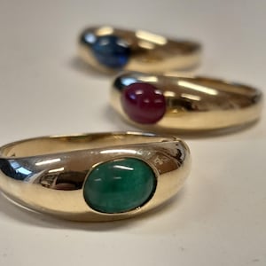 Op de afbeelding: Drie gouden ringen met ovale edelstenen. Eén ring heeft een groene edelsteen, een andere een rode edelsteen en de derde een blauwe edelsteen. De ringen hebben een gepolijst oppervlak, de stenen zijn gezet.