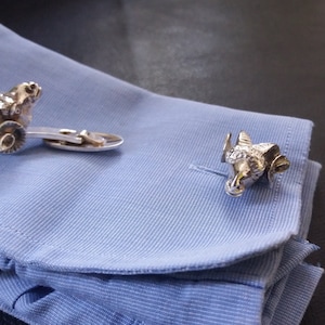 May include: A pair of silver cufflinks featuring a detailed ram's head design. The cufflinks are on a blue shirt cuff.