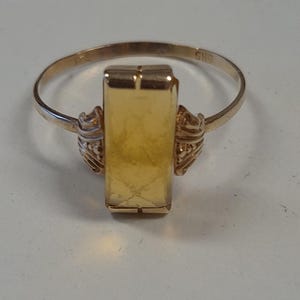 Op de afbeelding: Een gouden ring met een rechthoekige middensteen. De ring heeft decoratieve elementen aan de zijkanten van de middensteen en een dunne band. De ring ligt op een wit oppervlak.