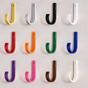 May include: A selection of twelve J-shaped wall hooks in a range of colours: yellow, white, black, red, orange, green, blue, purple, pink, brown, and off-white. These hooks are designed for hanging items and have a simple, modern design.